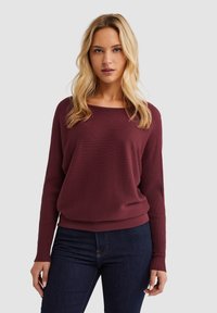 Bordeaux ribbeltrui met een relaxte pasvorm, brede halslijn en lange mouwen. Gecombineerd met donkerblauwe jeans, wat een casual stijl benadrukt.