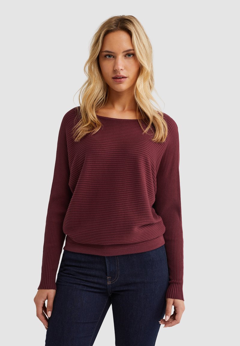 Bordeaux ribbeltrui met een relaxte pasvorm, brede halslijn en lange mouwen. Gecombineerd met donkerblauwe jeans, wat een casual stijl benadrukt.