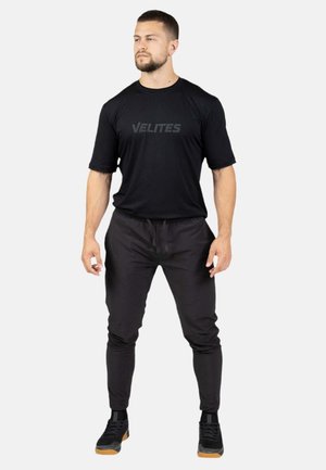 Hombre de pie con camiseta negra Velites, pantalones deportivos negros y zapatillas negras con suela de goma sobre un fondo liso y claro.