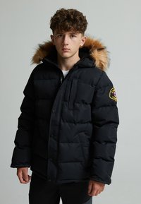 Black puffer jacket with a fur-lined hood, featuring a single chest pocket and a logo patch on the sleeve. Smooth, insulated fabric.
