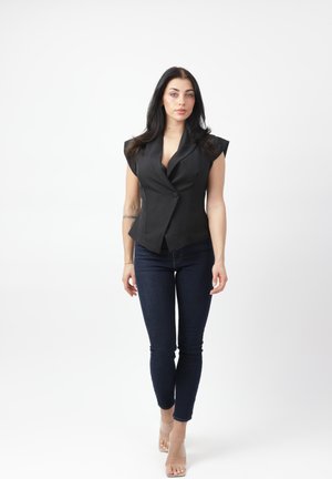 Kobieta o długich ciemnych włosach, ubrana w czarną bezrękawnikową marynarkę, ciemne obcisłe dżinsy i przezroczyste buty na wysokim obcasie, stoi na tle białego tła.