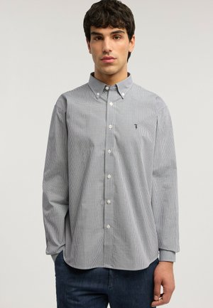 Mladý muž ve světlé košili s dlouhým rukávem a malým kostkovaným vzorem, zapnuté na knoflíky, a tmavých džínách, stojící proti jednoduchému světlému pozadí.