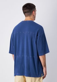 Niebieska,oversize'owa koszulka z okrągłym dekoltem, krótkimi rękawami i kontrastowymi szwami. Materiał wydaje się miękki i gładki w dotyku.