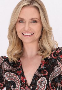 Blonďatá žena ve černých šatech s barevným paisley vzorem. Šaty mají hluboký výstřih ve tvaru V a nabrané balónové rukávy. Hladká textura.