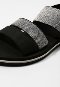 Musta sandaali, jossa on kolme leveää remmiä; kaksi on valmistettu tekstuurisesta hopeamateriaalista ja yksi mustasta kankaasta. Tyylissä on musta pohja, jossa on valkoisia yksityiskohtia.