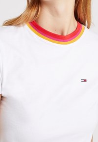 Camisa blanca con cuello acanalado en franjas amarillas, rosas y rojas, que muestra un pequeño logo de Tommy Hilfiger en el pecho.