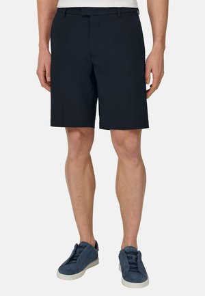 Uomo che indossa pantaloncini blu navy su misura con passanti per cintura e sneakers casual blu scuro su uno sfondo chiaro semplice.