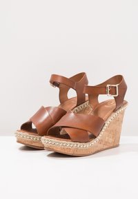 Sandali wedge in pelle marrone con cinturini incrociati, tacco in sughero e perline decorative lungo la suola. Chiusura con fibbia sul cinturino.