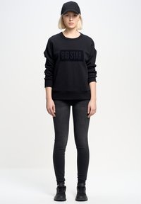 Crni oversized dukserica s "BIG STAR" logotipom, uparena s crnim skinny trapericama i crnim tenisicama, upotpunjena crnom kapom.