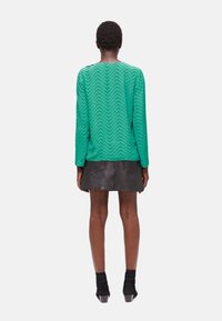 Green patterned long-sleeve top made of textured fabric, paired with a black leather skirt. Model wears black ankle boots.