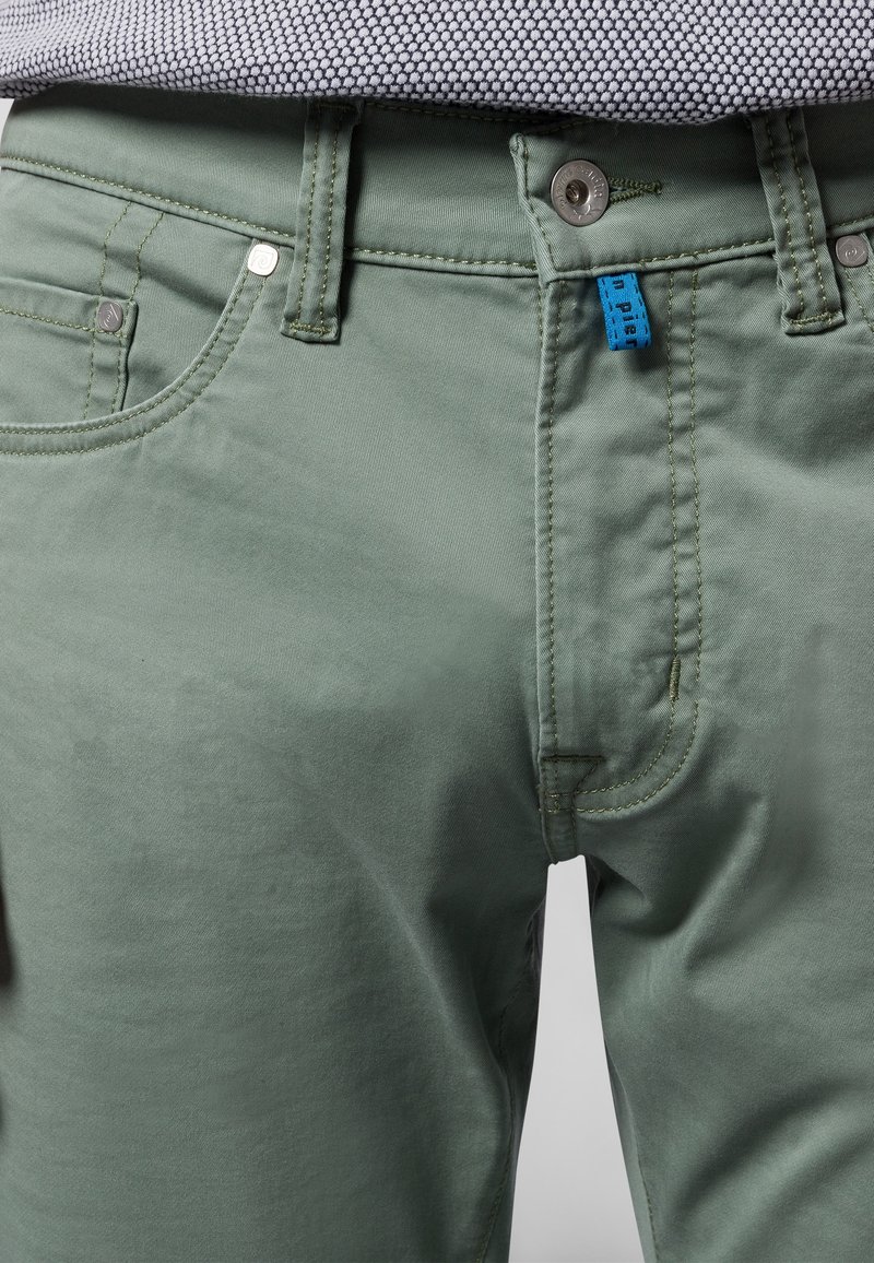 Nahaufnahme von grünen Hosen mit Fronttaschen, Gürtelschlaufen, einem Metallknopf und einem kleinen blauen Stoffetikett in der Nähe des Reißverschlusses.