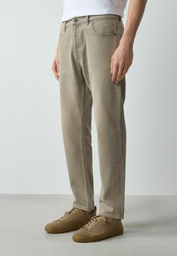 Mann trägt hellbraune Jeans und passende braune Wildledersneaker, steht auf einem glatten grauen Boden vor einer schlichten Wand.