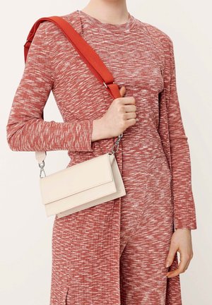 Persoon die een rood patroon outfit draagt en een kleine witte rechthoekige crossbodytas met een brede rode band over de schouder draagt.