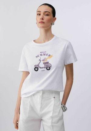 Donna che indossa una maglietta bianca con uno scooter viola e la scritta "Hello Summer", abbinata a pantaloni bianchi e orecchini a cerchio e braccialetti argentati.