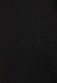 Tissu noir avec une texture lisse, arborant un logo "SMILODOX" brodé dans un fil assorti, positionné subtilement sur la surface.