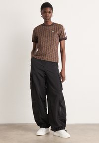 Braunes T-Shirt mit geometrischem Muster und schwarzem Saum, kombiniert mit schwarzen Cargo-Hosen mit Taschen und Reißverschlüssen, abgerundet mit weißen Sneakern.