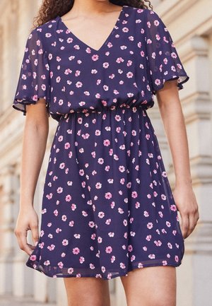Femme portant une robe bleu marine à imprimé floral rose et blanc, col en V, manches courtes volantées et taille cintrée, debout à l'extérieur.