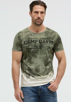 Mann trägt grünes und beiges kurzärmeliges T-Shirt mit schwarzem Text "Camp David" und blaue Jeans, steht vor neutralem hellem Hintergrund.