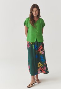 Top verde a maniche corte con bottoni abbinato a una lunga gonna verde scuro con motivi floreali colorati. Tessuto morbido, vestibilità casual.