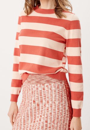 Kvinde iført en rød og cremefarvet bredstribet sweater parret med en rød og hvid tekstureret plisseret nederdel, stående mod en ensfarvet baggrund.