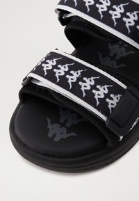 Černé sandály s nastavitelnými popruhy, které mají bílé opakující se siluety lidských postav a odpovídající logo tištěné na stélce.