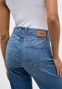 Blauwe denim jeans met een hoge taille, voorzien van een tan lederen patch met het label "ANGELS" en twee achterzakken met zichtbare stiksels.