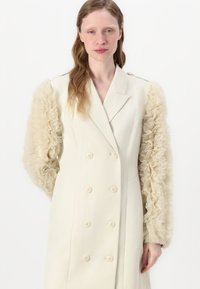 Manteau crème à double boutonnage avec des manches transparentes beiges à volants, présentant de grands boutons et un design ajusté. Texture lisse et forme structurée.
