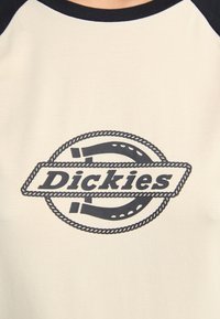Nahaufnahme eines hellen Hemdes mit einem runden Logo, auf dem "Dickies" in fetten Buchstaben steht, akzentuiert durch ein Seil-Design.