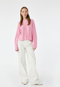 Maglione rosa a coste, oversize, con maniche lunghe, abbinato a pantaloni bianchi a gamba larga e scarpe chiare. Design semplice; nessun motivo o accento visibile.