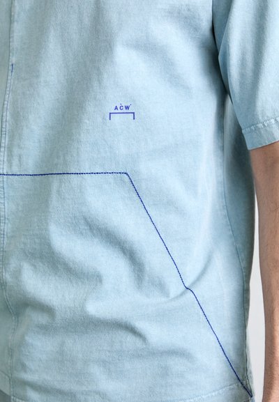 Chemise bleu clair en tissu doux, avec un logo violet et des coutures bleues le long des coutures et de la poche. Manches courtes et coupe décontractée.
