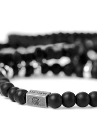 Schwarzes Perlenarmband mit mattschwarzen Perlen und einem rechteckigen grauen Metallelement, das mit einem Logo graviert ist. Glatte Oberfläche, schlichtes Design.