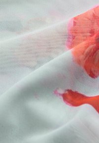 Biała siateczkowa tkanina z drobną teksturą siatki częściowo zakrywająca wzór czerwono-pomarańczowych kwiatów pod spodem.