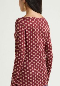 Langärmliges, kastanienbraunes Oberteil mit einem geometrischen Muster aus goldfarbenen und pinken Quadraten, gefertigt aus einem glatten, elastischen Stoff.