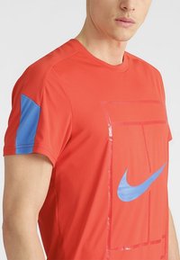 Röd tränings t-shirt med blå accent på vänster ärm. Har en blank Nike-logotyp och en geometrisk transparent design på framsidan.