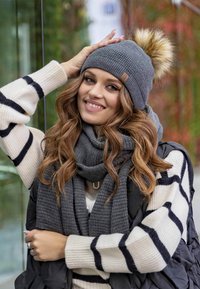 Grijze gebreide beanie met een bontpompon, gecombineerd met een bijpassende sjaal, gedragen over een crèmekleurige trui met zwarte strepen en een zwarte puffer vest.