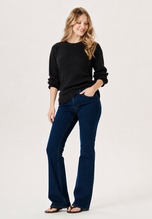 Young woman smiling, wearing black long-sleeve sweater, blue flared jeans, and sandals, standing with one hand in pocket.