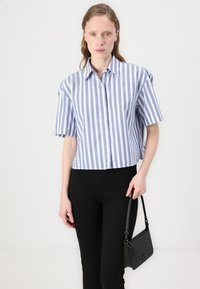 Blauw-wit gestreept overhemd met korte mouwen en een kraag, voorzien van een klein logo. Model houdt een zwarte handtas vast en draagt zwarte broek.