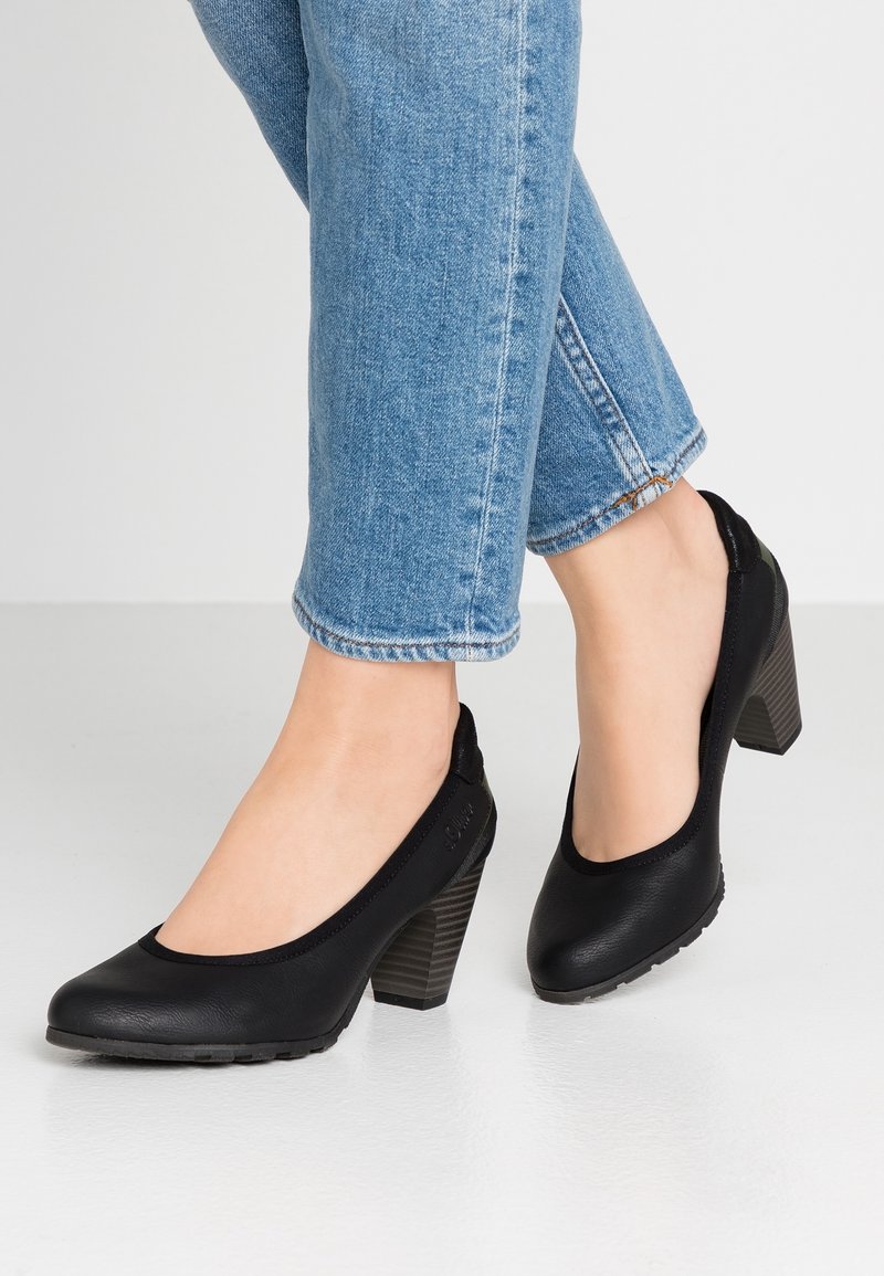Escarpins en cuir noir avec un bout arrondi, dotés d'un talon texturé empilé et de côtés élastiqués pour le confort. Portés avec un jean bleu raccourci.