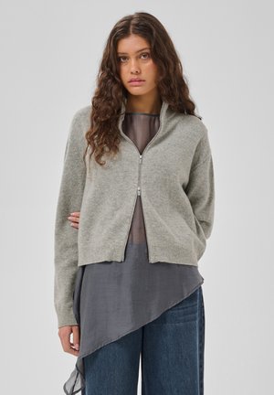 Femme aux longs cheveux ondulés portant un pull zippé gris clair sur un haut asymétrique gris foncé et un jean bleu, debout devant un fond uni.