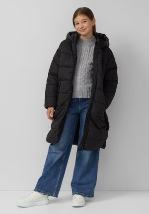 Meisje in een lange zwarte pufferjas, een grijze kabelgebreide trui, wijdvallende blauwe jeans en witte sneakers, staand tegen een eenvoudige muur.