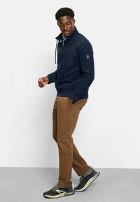 Marineblauer Fleece-Pullover mit Reißverschlusskragen und Brusttasche, kombiniert mit beigefarbenen Chinos und schwarz-grünen Sneakers. Bequemes, lässiges Design.