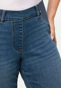 Jeans de denim en azul medio con una cintura alta, que cuentan con una banda elástica, costuras frontales y costuras naranjas en los bolsillos.