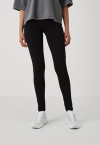 Leggings noirs en tissu extensible, ajustés, avec une texture lisse. Associés à un haut gris ample et des baskets blanches.