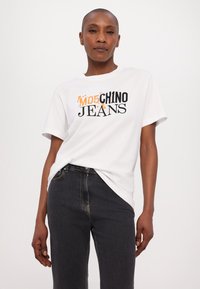 Bílá bavlněná tričko s černým a oranžovým grafickým textem "MO5 CHINO JEANS." Spárováno s tmavými džínami s přiléhavým střihem.