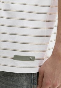 Camisa blanca de manga corta con rayas horizontales beige y una etiqueta verde en el dobladillo, hecha de un tejido suave con una textura lisa.