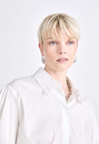 Camisa blanca de botones con cuello clásico, fabricada en un tejido suave y con un corte relajado. El modelo tiene el pelo rubio corto y lleva pendientes de aro plateados.