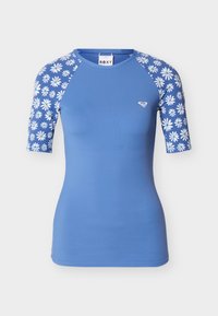 Μπλε ριπλέ ζιβάγκο με μοτίβα λευκής μαργαρίτας στα μανίκια και μικρό λευκό λογότυπο στο αριστερό στήθος.