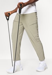 Light khaki cargo pants with multiple pockets, tapered design, and soft texture, paired with white athletic shoes and trekking poles.