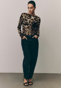 Maglietta a maniche lunghe con stampa leopardata trasparente in marrone e nero, abbinata a jeans scuri a vita alta e tacchi neri con cinturini, mani in tasca.