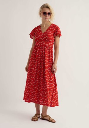 Nő piros virágos midi ruhában, barna szandálban, piros napszemüvegben és arany ékszerekben, egyszínű világos háttér előtt áll.